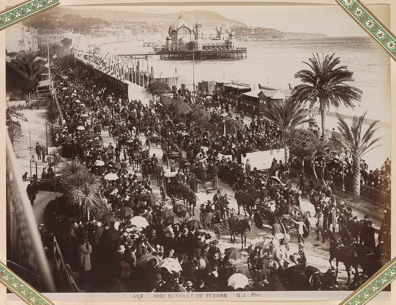 Cortège avec des spectateurs lors de la Bataille de fleurs à Nice ; une jetée avec des bâtiments en arrière-plan (circa 1900).